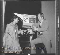 A woman with dark hair and a man in a light suit lean against a television camera as they have a discussion.