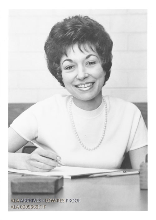 A young woman with short dark hair and a white shirt holds a pen over a pad of paper and smiles towards the camera.