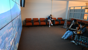 Two students watching a video in the Visualization Theatre while another studies in the background. The room consists of large monitors creating a singular screen, many rolling chairs, speakers for the monitor screen, and a charging statnion.