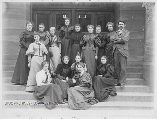 photo from the University of Illinois archives of 15 students posing for a photo in front of a building wearing attire from the late 1800s