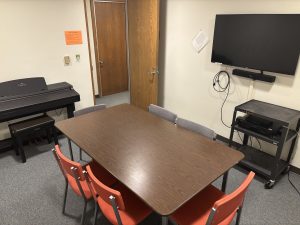 Room with table and six chairs, piano with bench, wall mounted screen and bluetooth soundbar, and cart with video playback equipment. 